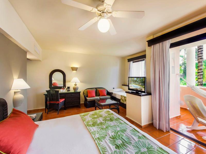 Modern hotel room with double bed, TV, and balcony overlooking palm trees.