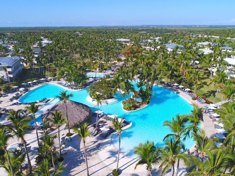 Large hotel pool with palm trees and umbrellas in a tropical resort under clear sky.