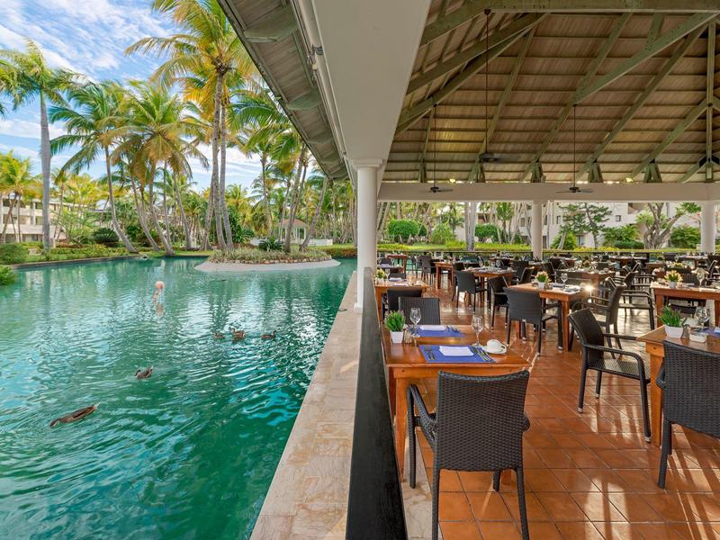 Outdoor restaurant with tables beside a large water feature and palm trees in the background.