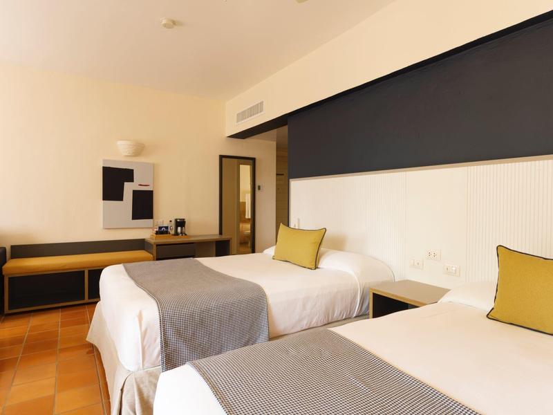 Hotel room with two single beds, gray throws, and yellow pillows, simply furnished.