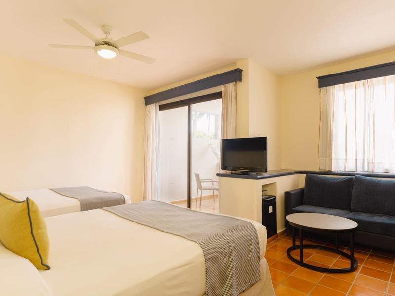 Bright hotel room with double bed, sofa, TV, and balcony access with seating area.