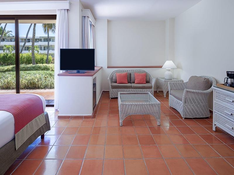 Bright hotel room with terracotta floor, sofas, TV, and garden view.