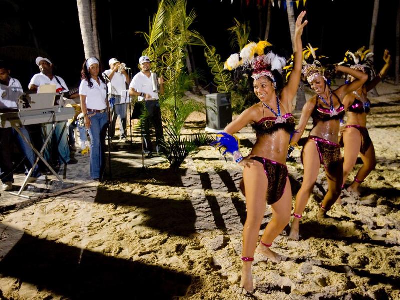 Three samba dancers perform at night on the beach, accompanied by musicians in the background.