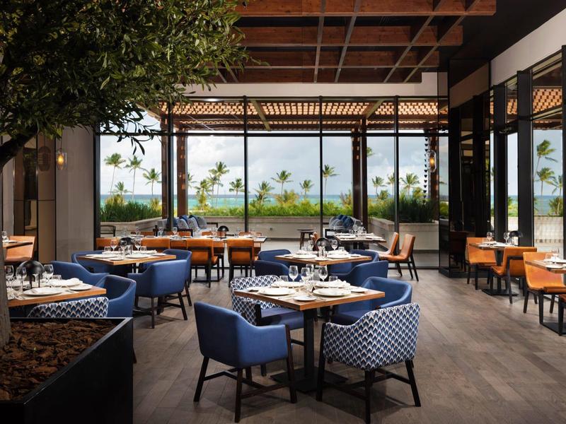 Restaurant moderne avec fenêtres du sol au plafond, chaises bleues, tables en bois et vue sur la nature.