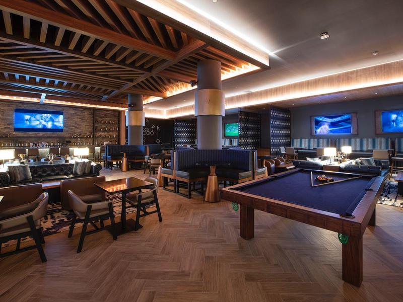 Espace lounge d'hôtel moderne avec table de billard, groupes de sièges et poutres en bois éclairées.