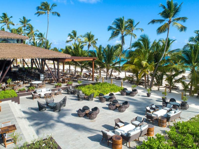 Espace de seating ouvert avec tables et chaises près des palmiers et plage en arrière-plan.