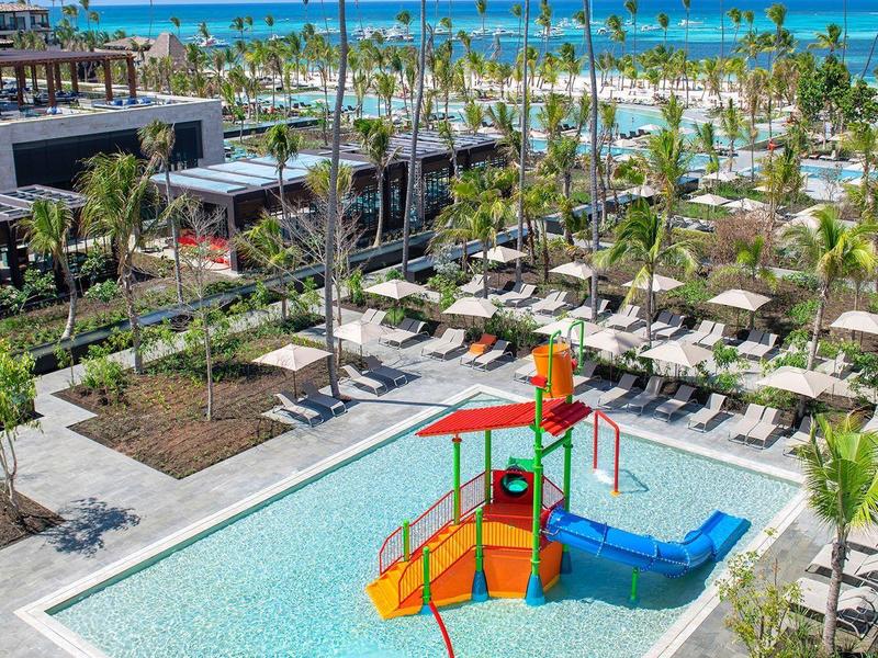 Piscine pour enfants avec aire de jeux colorée, entourée de chaises longues et de palmiers près de la plage.