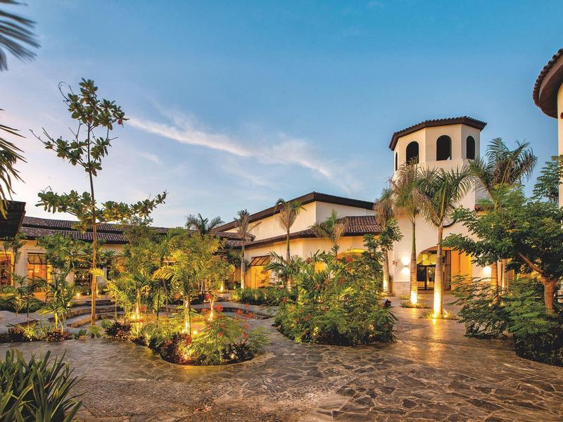 Hôtel luxueux de style méditerranéen, entouré d'une végétation luxuriante et d'une cour pavée.