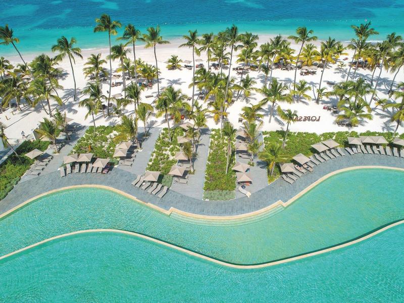 Une plage luxueuse avec du sable blanc, des palmiers et une piscine incurvée au bord de la mer turquoise.