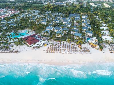 Luftaufnahme eines Strandes mit klarem Wasser, Liegestühlen, Palmen und Resortgebäuden dahinter.