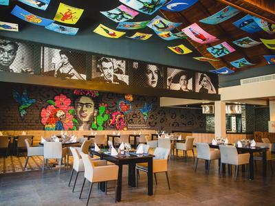 Modernes Restaurant mit bunter Deckenkunst und Wandporträts, hellen Stühlen und großen Tischen.