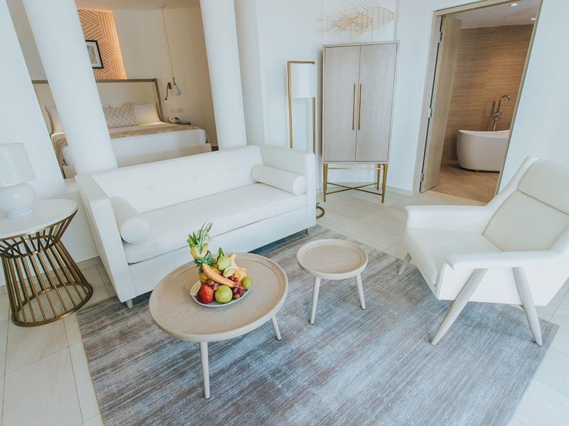 Sala luminosa con sofá blanco, sillón, dos mesas redondas y decoración de frutas sobre alfombra.