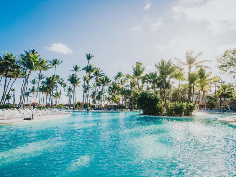 Gran piscina con agua clara rodeada de palmeras y cielo azul en un hotel tropical.