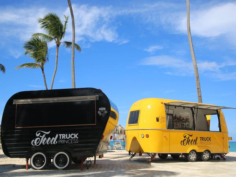 Dos food trucks, uno negro y otro amarillo, están en un área arenosa con palmeras al fondo.