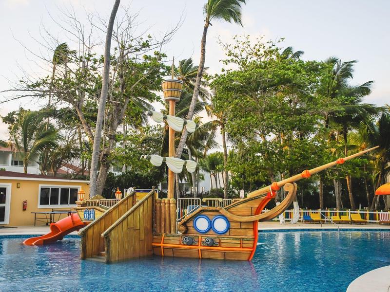 Parque acuático con barco pirata y tobogán en una piscina rodeada de palmeras y edificios.
