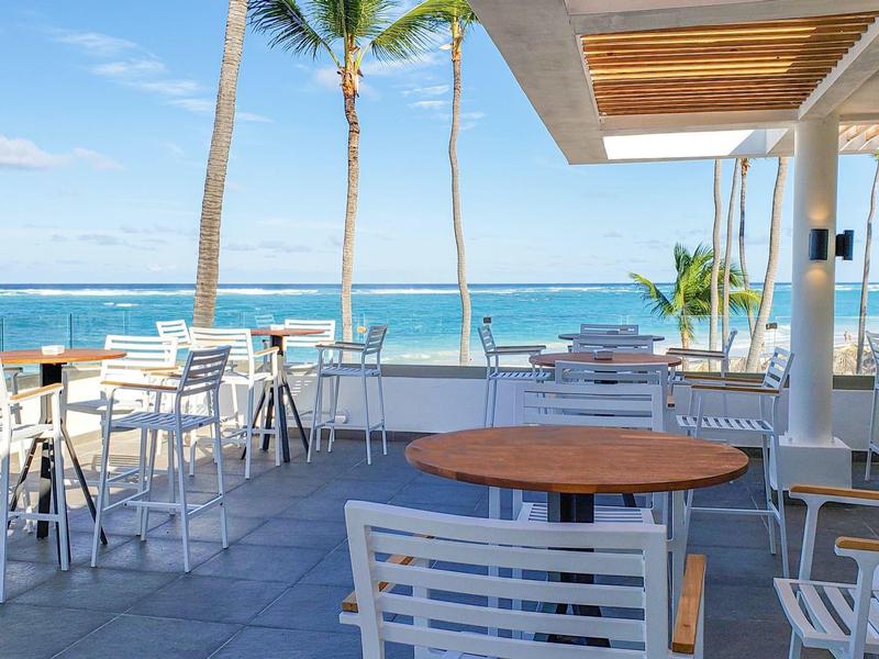 Terraza de hotel con mesas y sillas, vista al mar y palmeras bajo cielo despejado.