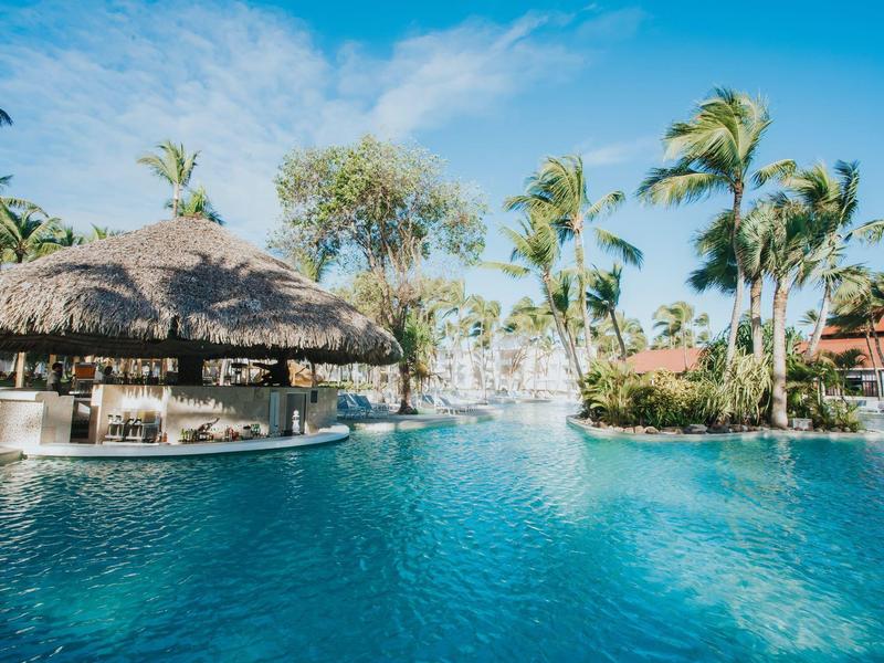 Gran piscina con bar swim-up bajo palmeras y cielo soleado en un complejo hotelero tropical.