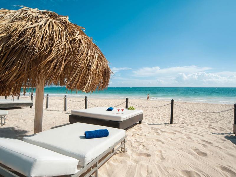 Playa con tumbonas blancas, sombrilla de paja y cielo azul sobre el mar.