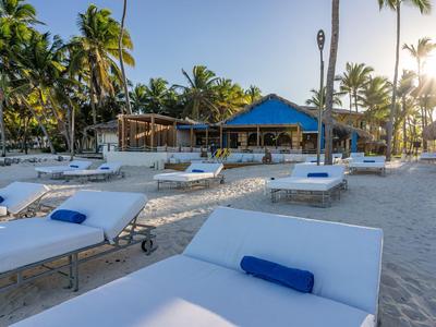 Strand mit Liegen und blauen Kissen vor einer Bar unter Palmen bei Sonnenuntergang.