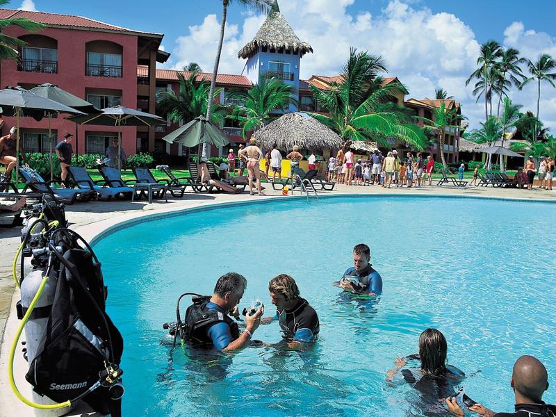 Persone con attrezzatura subacquea che imparano in una piscina d'hotel circondata da edifici tropicali e palme.