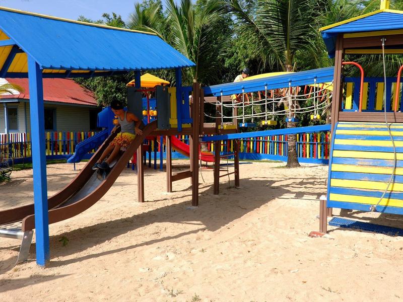 Area giochi colorata con scivoli e strutture per arrampicata su terreno sabbioso sotto palme.