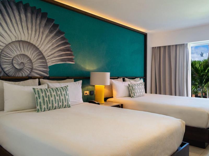 Camera d'hotel moderna con due letti, decorazione murale verde e balcone con vista sulle palme.
