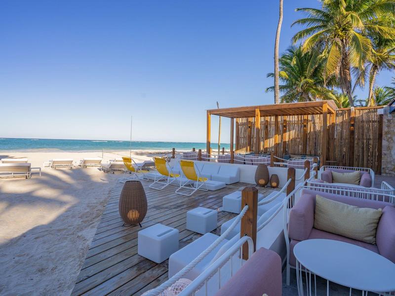 Lounge sulla spiaggia con divani bianchi, pavimento in legno e palme sotto il cielo azzurro