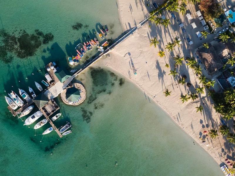 Luchtfoto van een strand met helder water, boten aan een steiger en palmbomen langs de kust.