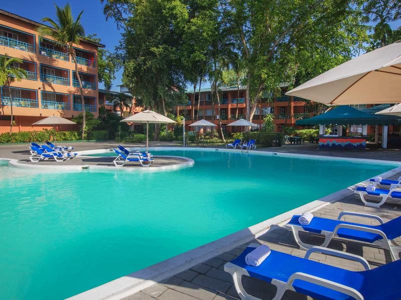Groot zwembad met blauwe ligstoelen en parasols voor een hotelgebouw met bomen.