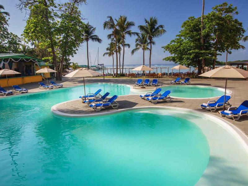 Rond hotelzwembad met blauwe ligstoelen en palmbomen op de achtergrond bij het strand.