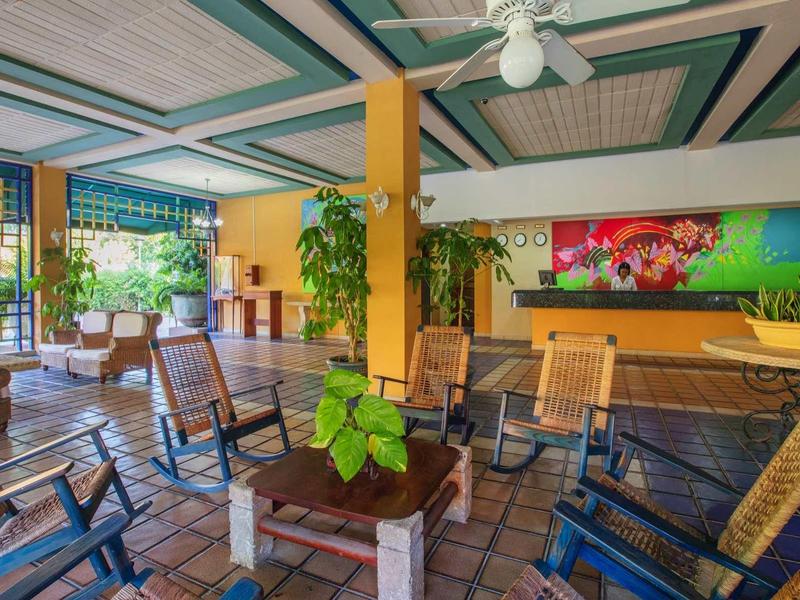 Ruime zithoek in hotel met houten meubels, planten en plafondventilator in een lichte kamer.