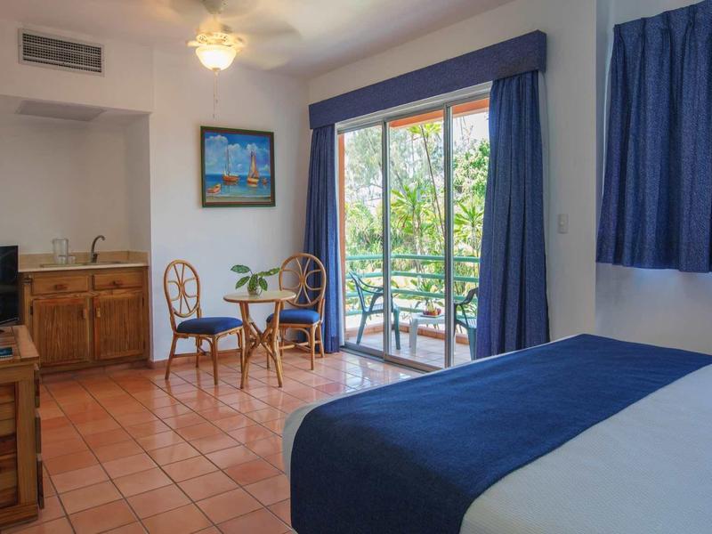 Lichte hotelkamer met tweepersoonsbed, tafel, stoelen en balkon met uitzicht op bomen.