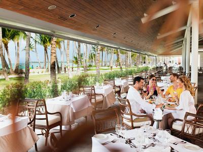 Restaurante al aire libre con huéspedes comiendo en mesas con manteles blancos bajo palmeras.