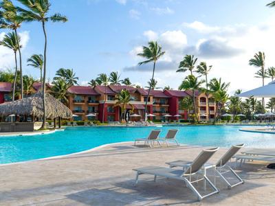 Zona piscina con lettini e ombrelloni davanti a un resort con palme.
