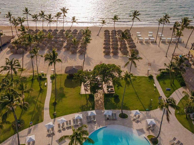 Uitzicht op een resortzwembad, tuin en strand met palmbomen en ligstoelen.