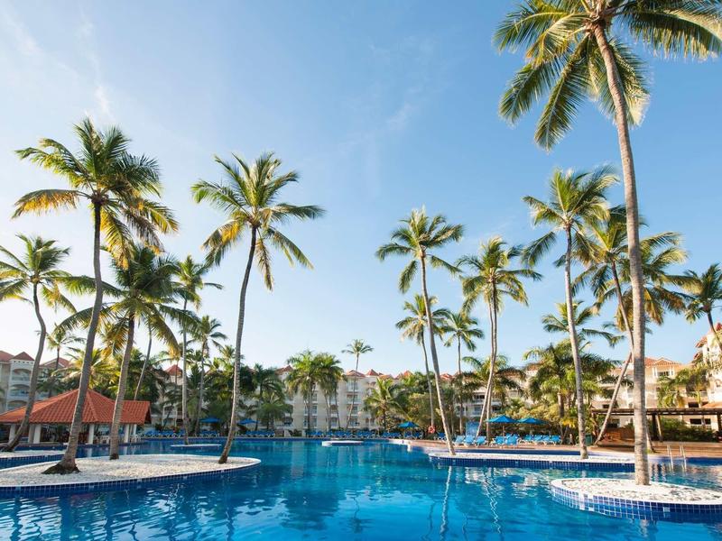 Large pool with umbrellas and palm trees at tropical resort under blue sky.