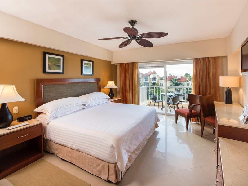 Bright hotel room with king-size bed, ceiling fan, and balcony seating with view.