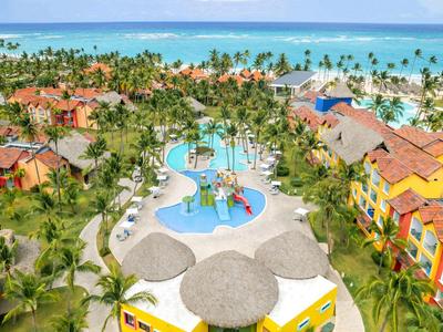 Hotel mit Pools und Palmen vor blauem Meer und Himmel an tropischem Urlaubsort.