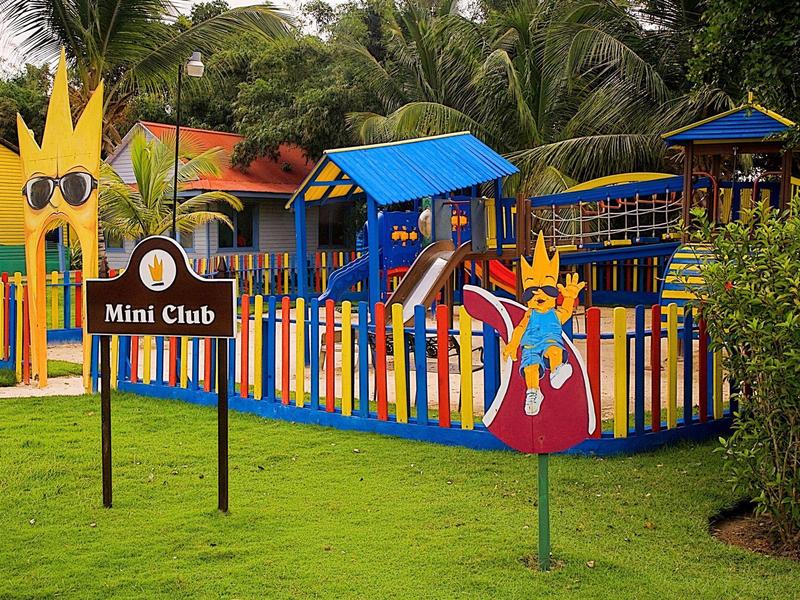 Kleurrijke speeltuin met hek, glijbanen en Mini Club bord in tropische omgeving.