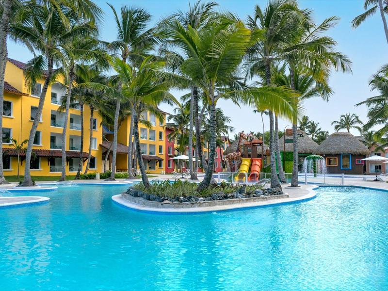 Groot hotelzwembad met palmbomen en geel gebouw op de achtergrond bij zonnig weer.