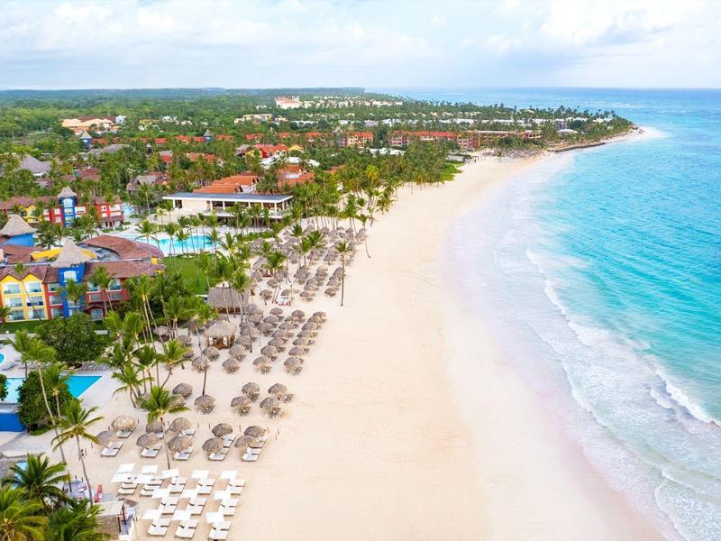 Lange witzandige strand met parasols, omgeven door tropische bomen en resorts.