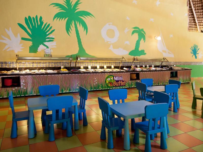 Chaises et tables colorées pour enfants devant un snack avec peinture murale tropicale.