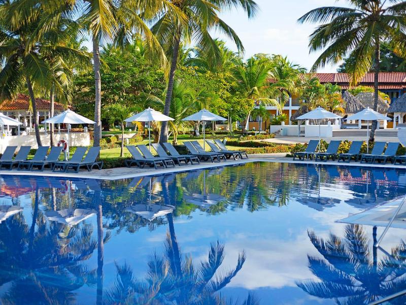 Grand piscine entourée de chaises longues, parasols et palmiers dans un hôtel tropical.
