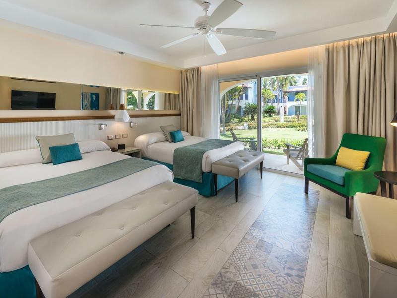 Chambre d'hôtel lumineuse avec deux lits, fauteuil vert et grandes fenêtres donnant sur le jardin.