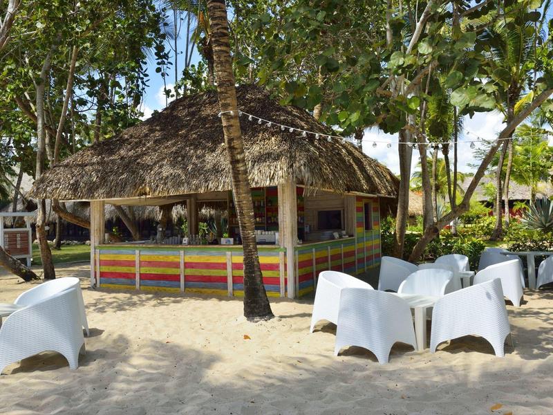 Bar de plage au toit de chaume entourée de palmiers et de chaises blanches sur le sable.