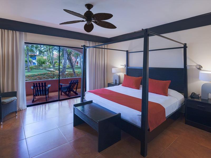 Chambre d'hôtel moderne avec lit à baldaquin, chaises sur la terrasse et vue sur le jardin.