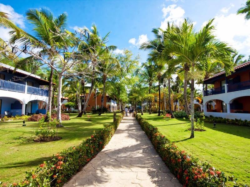 Allée bordée de palmiers et d'herbe menant à des bâtiments de villégiature sous un ciel bleu.