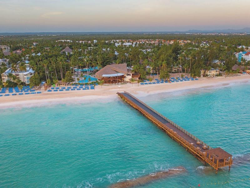 Langer hölzerner Steg ins türkisfarbene Meer mit weißem Sandstrand und grüner Küstenvegetation.