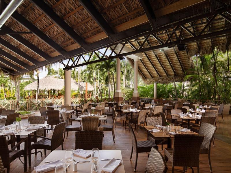 Offene Terrasse mit braunen Stühlen und Tischen, Holzdecke und grünen Pflanzen im Hintergrund.