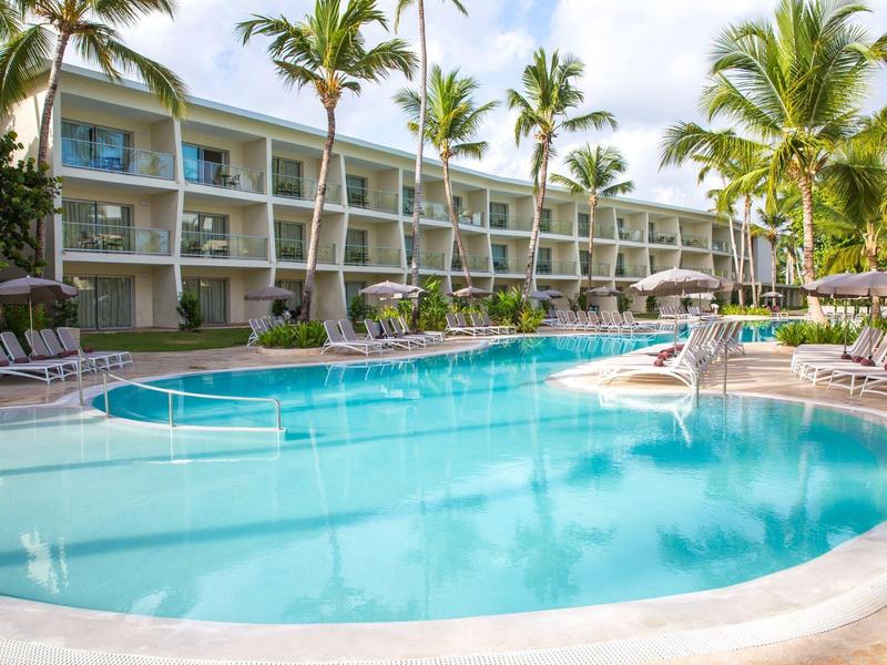 Großer Pool vor einem dreistöckigen Hotel mit Palmen und Liegestühlen bei bewölktem Himmel.